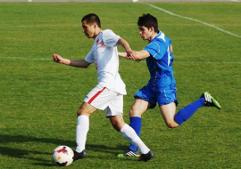 Moment intens dintr-un meci de fotbal cu doi jucători în acțiune pe teren. Un jucător în echipament alb controlează mingea, în timp ce adversarul în echipament albastru încearcă să-l oprească.